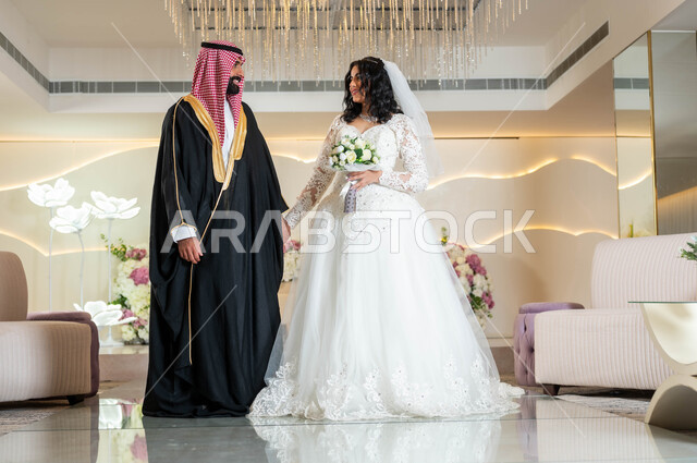 مفهوم الوفاء بالوعود، السرور والفرح في يوم الزواج، مراسم الأعراس والمناسبات في المملكة، صورة لعروس و عريس عربيان خليجيان سعوديان يمسكان الايدي ينظران لبعضهما بإيماءات الحب، قاعات الأفراح المزينة بالورود و الزهور، الاستقرار وتكوين عائلة