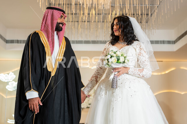 السرور والفرح في يوم الزواج، مراسم الأعراس والمناسبات في المملكة، صورة لعروس و عريس عربيان خليجيان سعوديان يمسكان الايدي ينظران لبعضهما بإيماءات الحب، مفهوم الوفاء بالوعود، قاعات الأفراح المزينة بالورود و الزهور، الاستقرار وتكوين عائلة