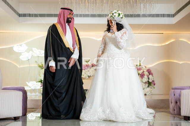 عريس عربي وسيم يرتدي الزي الرسمي والبشت التقليدي، مفهوم الخجل والحياء عند بنات العرب، عروسة عربية خليجية سعودية جميلة ترتدي فستان زفاف أبيض تقوم بتغطية وجهها بباقة الورد، أصالة العادات والتقاليد، مراسم الاعراس و المناسبات في المملكة، الفرح و السرور في يوم الزواج
