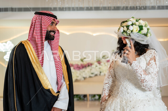 مفهوم الاناقة والاهتمام بالمظهر الخارجي، عريس يرتدي الزي الرسمي والبشت التقليدي، الخجل والحياء عند بنات العرب، عروسة عربية خليجية سعودية جميلة ترتدي فستان زفاف أبيض تقوم بتغطية وجهها بباقة الورد، مراسم الاعراس والمناسبات في المملكة، الفرح والسرور في يوم الزواج