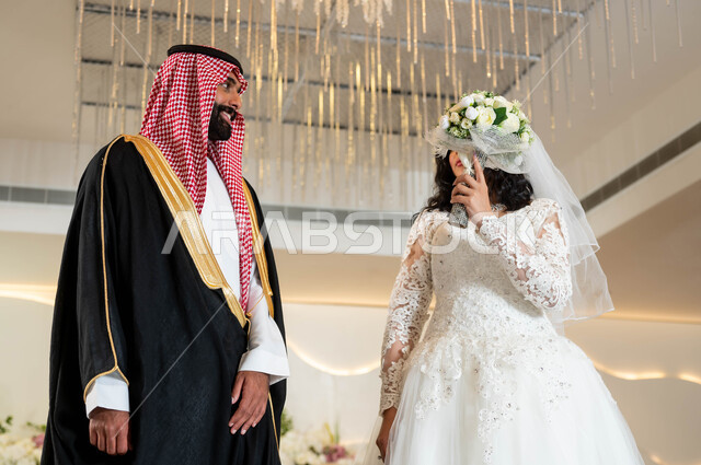 الاهتمام بالمظهر الخارجي، عريس عربي وسيم يرتدي الزي الرسمي والبشت التقليدي، مفهوم الخجل والحياء عند بنات العرب، عروسة عربية خليجية سعودية جميلة ترتدي فستان زفاف أبيض تقوم بتغطية وجهها بباقة الورد، مراسم الاعراس و المناسبات في المملكة، الفرح والسرور في يوم الزواج