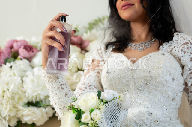 عطور ذات روائح فاخرة وفريدة، صورة مقربة لعروس عربية سعودية خليجيةترتدي ثوب زفاف أبيض تحمل زجاجة عطرية تقوم بوضع العطر، الاحتفال في قاعة المناسبات والأفراح, مراسم الأعراس والاحتفالات في المملكة، الفرح و السرورفي يوم الزواج