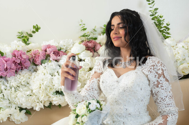 التطيب و التعطر بعطور ذات روائح فاخرة وفريدة، صورة مقربة لعروس عربية سعودية خليجيةترتدي ثوب زفاف أبيض تحمل زجاجة عطرية تقوم بوضع العطر، الاحتفال في قاعة المناسبات والأفراح, مراسم الأعراس والاحتفالات في المملكة، الفرح و السرورفي يوم الزواج