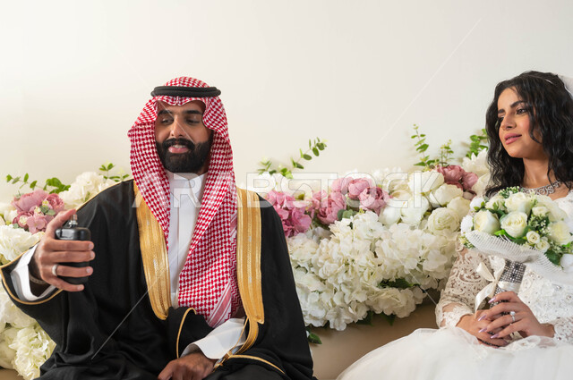 مفهوم الاناقة والاهتمام بالمظهر الخارجي، الفرح والسرور في يوم الزفاف، صورة مقربة لرجل عربي يرتدي الزي الرسمي والبشت التقليدي يقوم بوضع العطر، أجمل اطلالة لعروسة عربية خليجية سعودية ترتدي فستان زفاف أبيض تمسك بيدها باقة ورد، مراسم الاعراس والمناسبات في المملكة