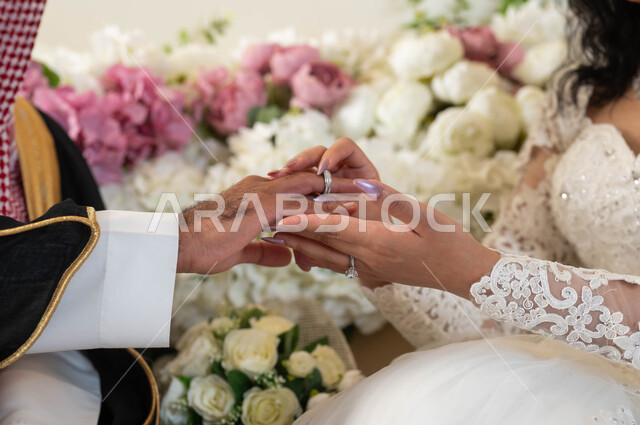 مراسم الاعراس والمناسبات في المملكة، الفرح والسرور في يوم الزفاف، استبدال خواتم الزواج، رجل عربي يرتدي الزي الرسمي والبشت التقليدي، صورة مقربة لعروسة عربية خليجية سعودية ترتدي فستان أبيض تقوم بوضع الخاتم للمعرس، صالة الافراح و المناسبات