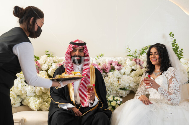 عريس وسيم عربي يرتدي الزي الرسمي البشت التقليدي، عروس جميلة ترتدي فستان زفاف أبيض تحمل بيدها كأس عصير، الفرح و السرور في يوم الزواج، مراسم الأعراس و المناسبات، مضيفة تقوم بتقديم الحلويات للعرسان، الضيافة في قاعات الأفراح والاحتفالات