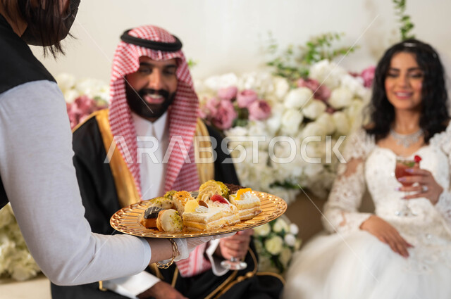 A hostess serving sweets to the grooms, hospitality in wedding halls and celebrations, a handsome Arab groom wearing the traditional Bisht uniform, a beautiful bride wearing a white wedding dress holding a glass of juice in her hand, joy and pleasure on the wedding day, wedding ceremonies and events.