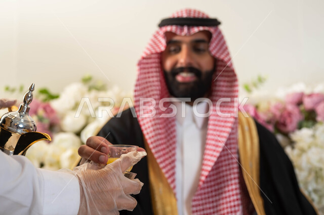 مراسم الأعراس والزواج في السعودية, صورة مقربة لمضيفة تقوم بتقديم القهوة السعودية الأصيلة، الاحتفال في قاعة المناسبات والأفراح, عريس عربي خليجي سعودي يرتدي الزي الرسمي والبشت التقليدي يأخذ فنجان المشروبات الساخنة, إيماءات السرور والفرح