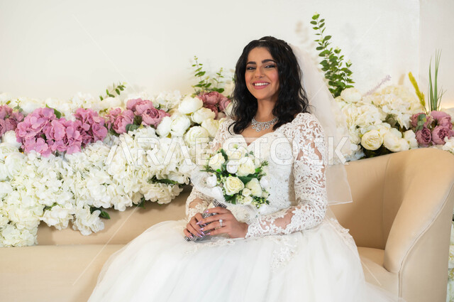 قاعات الأفراح و المناسبات المزينة بالزهور، مراسم الأعراس والاحتفالات في المملكة، الاهتمام بالاناقة و المظهر الخارجي، مفهوم التزين والاطلالة المتألقة، عروس عربية خليجية سعودية ترتدي فستان الزفاف الأبيض تمسك بيدها باقة ورد، الفرح و السرور في يوم الزواج