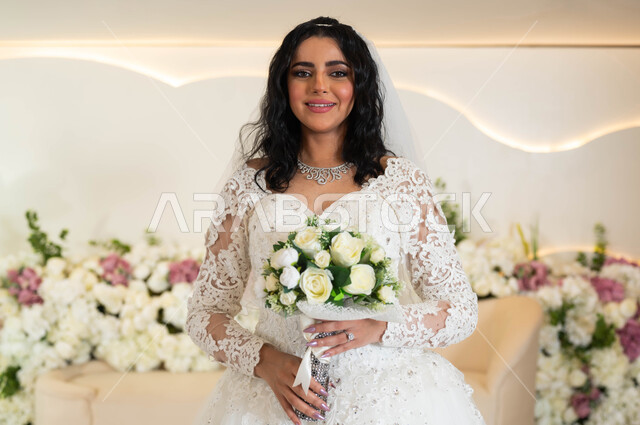 النظر إلى الكاميرا بإيماءات الفرح والسرور, مراسم الأعراس والزواج في السعودية، صورة مقربة لعروس عربية خليجية سعودية مبتسمة ترتدي فستان الزفاف الأبيض تمسك بيدها باقة ورد تقف في قاعة الأفراح و المناسبات المزينة بالزهور, مفهوم التزين والاطلالة المتألقة