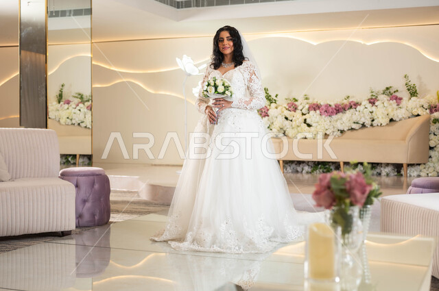 Attention to elegance and external appearance, wedding and marriage ceremonies in Saudi Arabia, the concept of adornment and a radiant appearance, a Saudi Arabian Gulf bride wearing a white wedding dress, holding a bouquet of roses in her hand, standing in the wedding and event hall decorated with flowers, full-length body imag