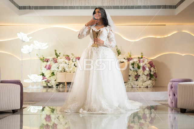 أصالة العادات والتقاليد السعودية، الاحتفال في قاعة المناسبات والأفراح, عروس عربية خليجية سعودية مبتسمة ترتدي فستان الزفاف الأبيض تحمل بيدها حامل البخور( المبخرة ) تقوم بتبخير نفسها، التطيب بالمسك والعود، مراسم الأعراس والزواج, صورة لكامل الجسم