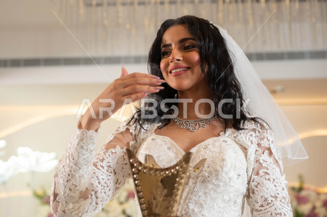 الاحتفال في قاعة المناسبات والأفراح, صورة مقربة لعروس عربية خليجية سعودية مبتسمة ترتدي فستان الزفاف الأبيض تحمل بيدها حامل البخور( المبخرة ) تقوم بتبخير نفسها، التطيب بالمسك والعود، أصالة العادات والتقاليد السعودية، مراسم الأعراس والزواج