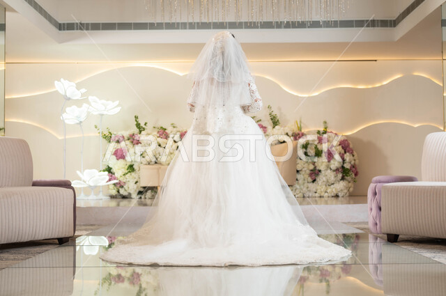 The concept of a sparkling look, attention to elegance and external appearance, a picture from the back of a Saudi Arabian Gulf bride wearing a white wedding dress standing in a wedding and event hall decorated with flowers, wedding and marriage ceremonies in Saudi Arabia, a full-length body photo