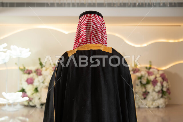 مفهوم الرجولة والأناقة والاهتمام بالمظهر الخارجي، صورة من الخلف لعريس عربي سعودي خليجي يرتدي الزي الرسمي والبشت التقليدي يقف في قاعة المناسبات والأفراح الفاخرة المزينة بالورود, مراسم الأعراس والزواج في السعودية