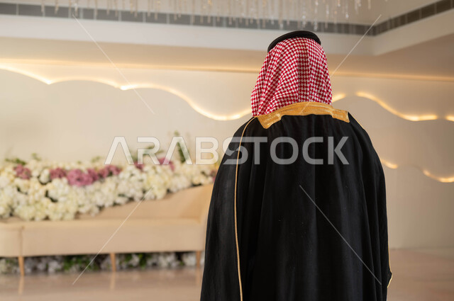 الاهتمام بالأناقة والمظهر الخارجي، صورة من الخلف لعريس عربي سعودي خليجي يرتدي الزي الرسمي والبشت التقليدي يقف في قاعة المناسبات والأفراح الفاخرة المزينة بالورود, مراسم الأعراس والزواج في السعودية