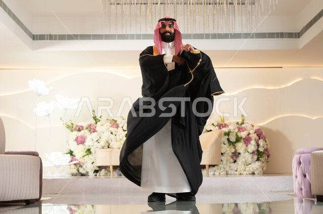 Celebrating in an event and wedding hall decorated with flowers, attention to elegance and external appearance, a Saudi Arabian Gulf groom wearing formal attire and a traditional bisht, standing straight and looking at the camera with gestures of happiness and joy, wedding and marriage ceremonies in Saudi Arabia, full-length body phot