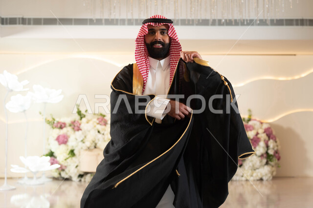 الأناقة والاهتمام بالمظهر الخارجي، عريس عربي سعودي خليجي يرتدي الزي الرسمي والبشت التقليدي يقوم بتعديل ثوبه ينظر إلى الكاميرا بإيماءات السعادة والفرح، الاحتفال في قاعة المناسبات والأفراح المزينة بالورود, مراسم الأعراس والزواج في السعودية