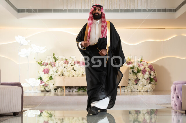 The concept of elegance and attention to external appearance, a Saudi Arabian Gulf groom wearing formal attire and a traditional bisht, standing straight and looking at the camera with joyful gestures, celebrating in an event and wedding hall decorated with flowers, wedding and marriage ceremonies in Saudi Arabia, full-length body photo