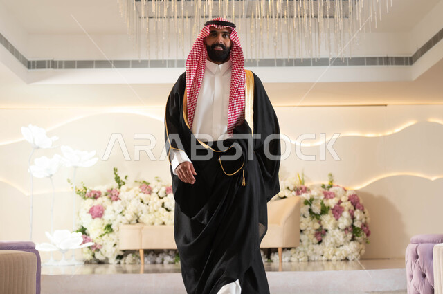 الاهتمام بالأناقة والمظهر الخارجي، عريس عربي سعودي خليجي يرتدي الزي الرسمي والبشت التقليدي يقف باستقامة ينظر إلى الكاميرا بإيماءات السعادة والفرح، الاحتفال في قاعة المناسبات والأفراح المزينة بالورود, مراسم الأعراس والزواج في السعودية
