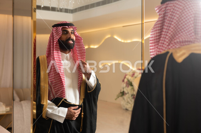 الاهتمام بالأناقة والمظهر الخارجي، صورة من الخلف لعريس عربي سعودي خليجي يرتدي الزي الرسمي والبشت التقليدي يقف أمام المرآة يقوم بتعديل ثوبه، إيماءات السعادة والفرح، الاستعداد للاحتفال في قاعة الأفراح والمناسبات, مراسم الأعراس والزواج في السعودية