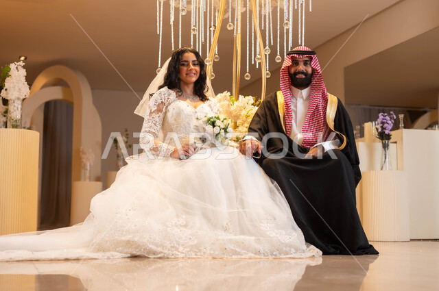 النظر إلى الكاميرا بإيماءات الحب والفرح، مفهوم الإرتباط وتكوين الأسرة، رجل يرتدي الزي الرسمي والبشت التقليدي، عروس عربية خليجية سعودية ترتدي فستان الزفاف الأبيض تمسك بيدها باقة الورود، الاحتفال في قاعة المناسبات والأفراح, مراسم الزواج والأعراس في السعودية