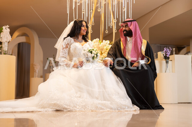 إيماءات الحب والفرح، مفهوم الإرتباط وتكوين الأسرة، رجل يرتدي الزي الرسمي والبشت التقليدي، عروس عربية خليجية سعودية ترتدي فستان الزفاف الأبيض تمسك بيدها باقة الورود تنظر إلى زوجها، الاحتفال في قاعة المناسبات والأفراح, مراسم الزواج والأعراس في السعودية