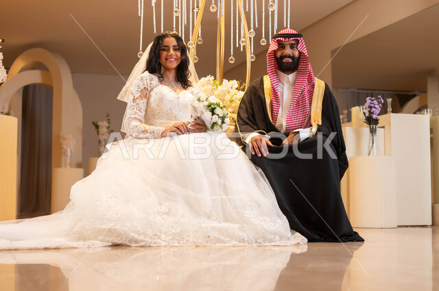 النظر إلى الكاميرا بإيماءات الفرح، مفهوم الإرتباط وتكوين الأسرة، رجل يرتدي الزي الرسمي والبشت التقليدي، عروس عربية خليجية سعودية ترتدي فستان الزفاف الأبيض تمسك بيدها باقة الورود، الاحتفال في قاعة المناسبات والأفراح, مراسم الأعراس والزواج في السعودية