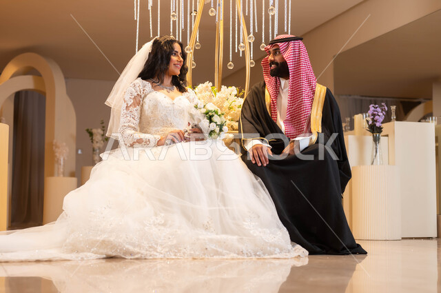 مفهوم الارتباط وتكوين الأسرة، رجل يرتدي الزي الرسمي والبشت التقليدي، عروس عربية خليجية سعودية ترتدي فستان الزفاف الأبيض تمسك بيدها باقة الورود، النظر إلى الكاميرا بإيماءات السرور، الاحتفال في قاعة المناسبات والأفراح, مراسم الزواج والأعراس في السعودية