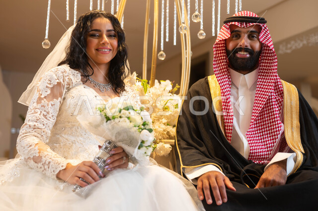 مفهوم الارتباط والألفة، رجل يرتدي الزي الرسمي والبشت التقليدي، عروس عربية خليجية سعودية ترتدي فستان الزفاف الأبيض تمسك بيدها باقة الورود، النظر إلى الكاميرا بإيماءات السرور، الاحتفال في قاعة المناسبات والأفراح, مراسم الأعراس والزواج في السعودية