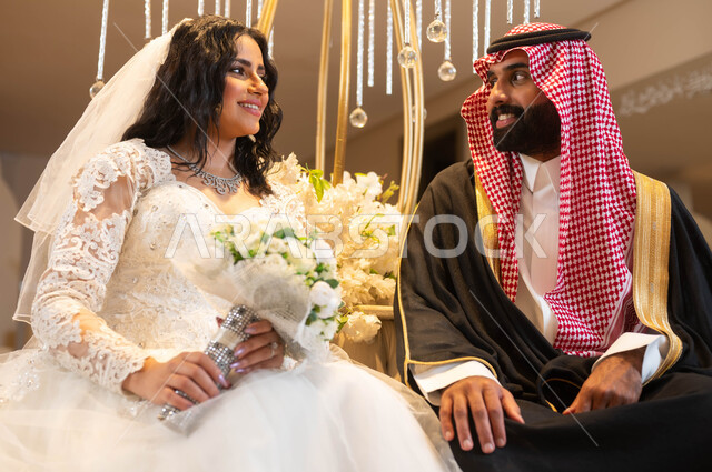 رجل يرتدي الزي الرسمي والبشت التقليدي، عروس عربية خليجية سعودية ترتدي فستان الزفاف الأبيض تمسك بيدها باقة الورود تنظر إلى العريس بإيماءات الحب، الاحتفال في قاعة المناسبات والأفراح المزينة بالأزهار ، مراسم الأعراس والزواج في السعودية