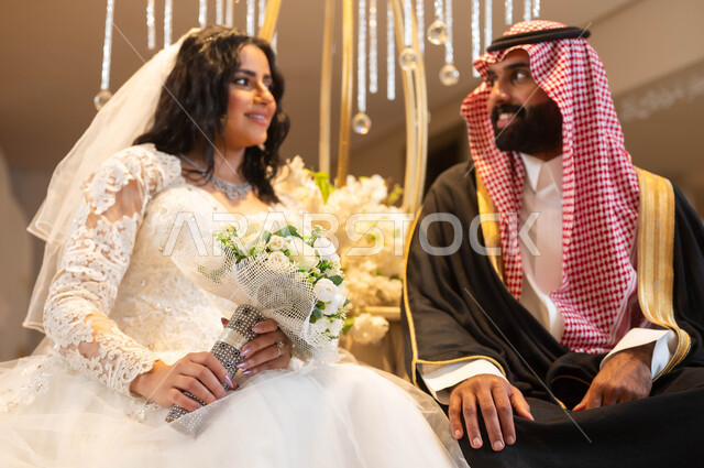 شاب يرتدي الزي الرسمي والبشت التقليدي، عروس عربية خليجية سعودية ترتدي فستان الزفاف الأبيض تمسك بيدها باقة الورود تنظر إلى العريس بإيماءات الحب، الاحتفال في قاعة المناسبات والأفراح المزينة بالأزهار ، مراسم الأعراس والزواج في السعودية