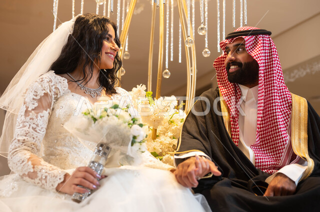 شاب يرتدي الزي الرسمي والبشت التقليدي، عروس عربية خليجية سعودية ترتدي فستان الزفاف الأبيض تمسك يد زوجها تنظر إليه بإيماءات السرور، الاحتفال في قاعة المناسبات والأفراح المزينة بالأزهار والورود، مراسم الأعراس والزواج في السعودية