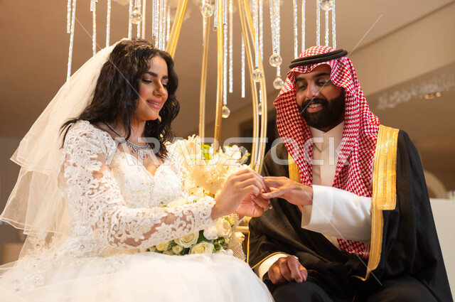 الاحتفال في قاعة المناسبات والأفراح المزينة بالأزهار والورود، رجل يرتدي الزي الرسمي والبشت التقليدي،عروس عربية خليجية سعودية ترتدي فستان الزفاف الأبيض تقوم بوضع خاتم الزواج بيد العريس، مراسم الأعراس والزواج في السعودية