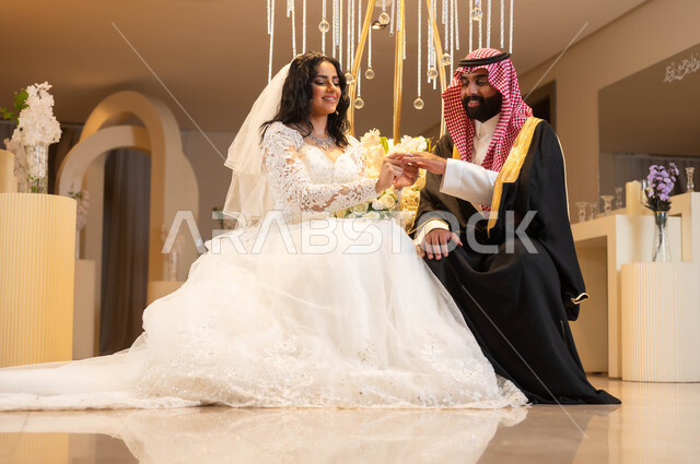 رجل يرتدي الزي الرسمي والبشت التقليدي، عروس عربية خليجية سعودية ترتدي فستان الزفاف الأبيض تقوم بوضع خاتم الزواج بيد العريس، مراسم الأعراس والزواج في السعودية، الاحتفال في قاعة المناسبات والأفراح المزينة بالأزهار والورود