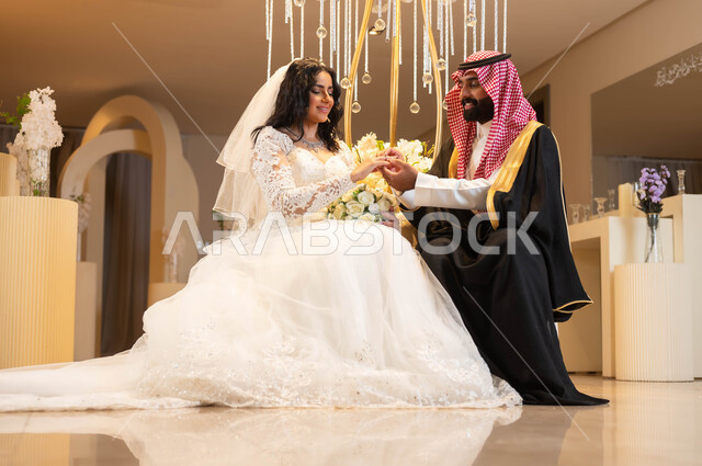 الاحتفال في قاعة المناسبات والأفراح المزينة بالأزهار والورود, امرأة ترتدي فستان الزفاف الأبيض، صورة مقربة لأيدي عريس عربي خليجي سعودي يرتدي الزي الرسمي والبشت يقوم بوضع خاتم الزواج بيد العروس، مراسم الأعراس والزواج في السعودية