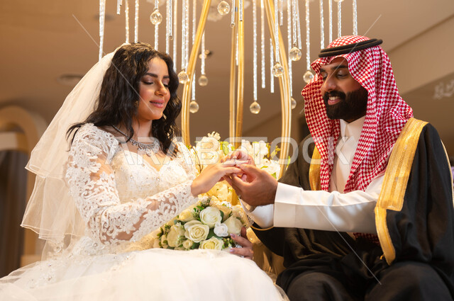 امرأة ترتدي فستان الزفاف الأبيض، صورة مقربة لأيدي عريس عربي خليجي سعودي يرتدي الزي الرسمي والبشت يقوم بوضع خاتم الزواج بيد العروس، مراسم الأعراس والزواج في السعودية ، الاحتفال في قاعة المناسبات والأفراح المزينة بالأزهار والورود
