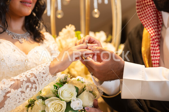 مراسم الأعراس والزواج في السعودية ، صورة مقربة لأيدي عريس عربي خليجي سعودي يرتدي الزي الرسمي والبشت يقوم بوضع خاتم الزواج بيد العروس، امرأة ترتدي فستان الزفاف الأبيض، الاحتفال في قاعة المناسبات والأفراح المزينة بالأزهار والورود