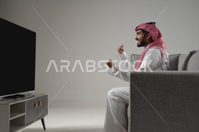 تعابير النصر والفوز, متابعة مباراة كرة القدم, التحدي والتشجيع والقوة, ايماءات الشغف والحماس بالانتصار, رفع اليدين الى الاعلى بتحقيق النجاح, بورتريه لشاب عربي سعودي خليجي يرتدي الثوب التقليدي والشماغ, قضاء اوقات ممتعة ومسلية, خلفية رمادية