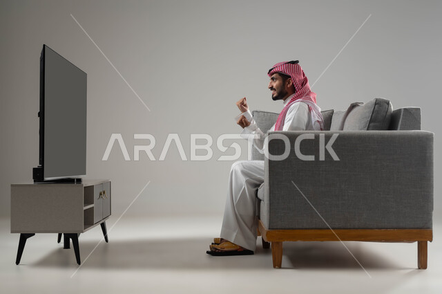 متابعة مباراة كرة القدم, الانسجام والتفاعل مع المنافسة, التحدي والتشجيع والقوة, ايماءات الشغف والحماس بالفوز, رفع اليدين الى الاعلى بتعابير الفوز, بورتريه لشاب عربي سعودي خليجي يرتدي الثوب التقليدي والشماغ, قضاء اوقات ممتعة ومسلية, خلفية رمادية