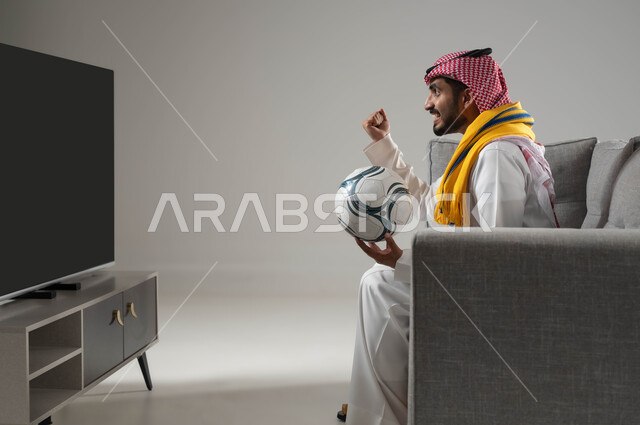 الشغف بمباريات كرة الفدم, الانسجام والتفاعل مع المنافسة, رفع اليد الى الاعلى بتعابير الفوز والانتصار, بورتريه لشاب عربي سعودي خليجي يرتدي الثوب التقليدي والشماغ يمسك الكرة, ارتداء وشاح التشجيع, خلفية رمادية