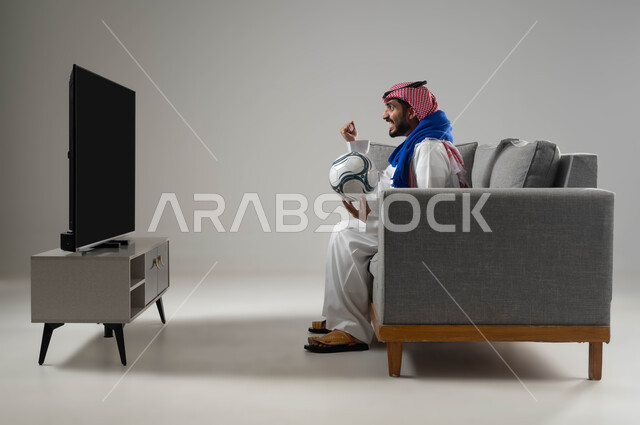 المنافسة والتحدي والتشجيع, النجاح والقوة وتحقيق الاهداف, ايماءات الشغف والحماس بالفوز, متابعة المباريات والالعاب الرياضية, الشغف بمباراة كرة القدم, بورتريه لشاب عربي سعودي خليجي يرتدي الثوب التقليدي والشماغ يمسك بيده الكرة, ارتداء وشاح تشجيعي, خلفية رمادية