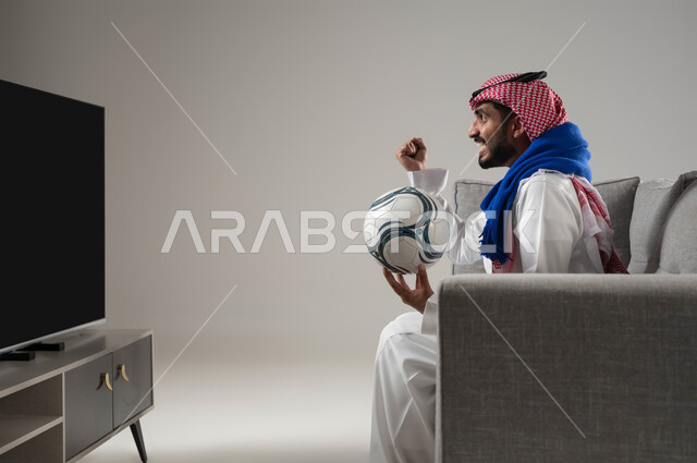 النجاح والقوة وتحقيق الاهداف, المنافسة والتحدي والتشجيع, ايماءات الشغف والحماس بالفوز, متابعة المباريات والالعاب الرياضية, الشغف بمباراة كرة القدم, بورتريه لشاب عربي سعودي خليجي يرتدي الثوب التقليدي والشماغ يمسك بيده الكرة, ارتداء وشاح تشجيعي, خلفية رمادية