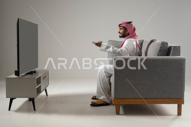 اختيار فيلم او برنامج للمشاهدة, استخدام التكنولوجيا في الترفيه, قضاء وقت الفراغ بمشاهدة الافلام المفضلة, نشاط منزلي ترفيهي فردي, بورتريه لشاب عربي سعودي خليجي يرتدي الثوب التقليدي والشماغ يجلس على الاريكة يمسك بيده جهاز التحكم, خلفية رمادية