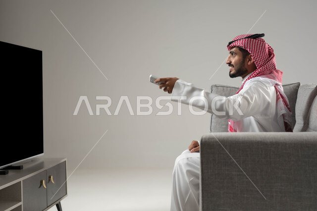 مشاهدة المسلسلات والبرامج التلفزيونية, استخدام التكنولوجيا في الترفيه, قضاء وقت الفراغ بمشاهدة الافلام المفضلة, نشاط منزلي ترفيهي فردي, بورتريه من الجانب لشاب عربي سعودي خليجي يرتدي الثوب التقليدي والشماغ يجلس على الاريكة يمسك بيده جهاز التحكم, خلفية رمادية