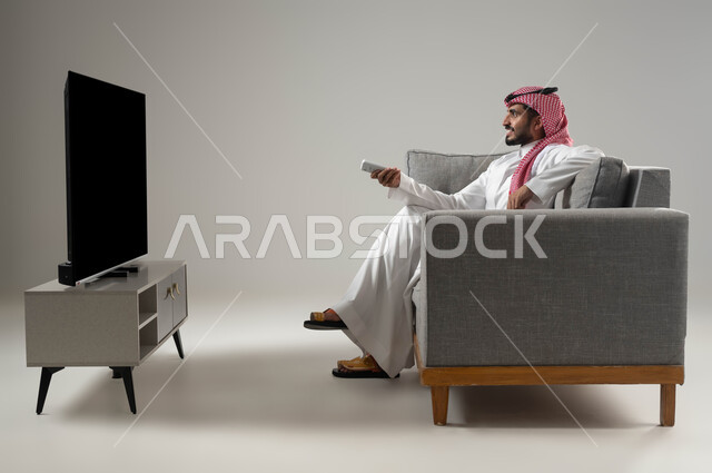 استخدام التكنولوجيا في الترفيه, قضاء وقت الفراغ بمشاهدة الافلام المفضلة, نشاط منزلي ترفيهي فردي, مشاهدة الافلام والمسلسلات, بورتريه لشاب عربي سعودي خليجي يرتدي الثوب التقليدي والشماغ يجلس على الاريكة يمسك بيده جهاز التحكم, خلفية رمادية