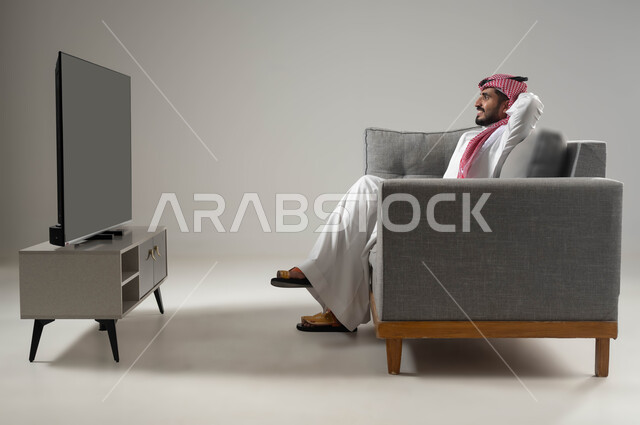 نشاط منزلي ترفيهي فردي, الاستجمام وقضاء اوقات ممتعة بمشاهدة التلفاز, ايماءات الراحة والاسترخاء, مشاهدة الافلام والمسلسلات, بورتريه لشاب عربي سعودي خليجي يجلس على الاريكة يضع يديه فوق رأسه, خلفية رمادية
