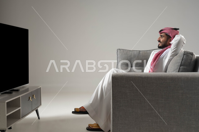 الاستجمام وقضاء اوقات ممتعة بمشاهدة التلفاز, نشاط منزلي ترفيهي فردي, ايماءات الراحة والاسترخاء, مشاهدة الافلام والمسلسلات, بورتريه لشاب عربي سعودي خليجي يجلس على الاريكة يضع يديه فوق رأسه, خلفية رمادية