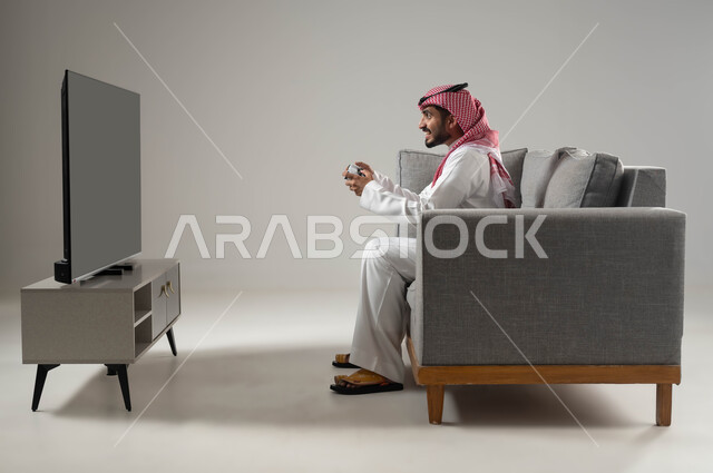 Using advanced modern technical devices, entertaining and enjoyable PlayStation games, the concept of winning and losing, spending an enjoyable time playing video games, a portrait of a Saudi Gulf Arab young man wearing a traditional thobe and shemagh, sitting on a comfortable sofa holding a joystick in his hand, gray background