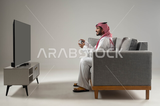العاب البلايستيشن الترفيهية والممتعة, مفهوم الفوز والخسارة, استخدام اجهزة تقنية حديثة متطورة, قضاء وقت ممتع بألعاب الفيديو, بورتيه لشاب عربي سعودي خليجي يرتدي الثوب التقليدي والشماغ يجلس على اريكة مريحة يمسك بيده عصا التحكم, خلفية رمادية